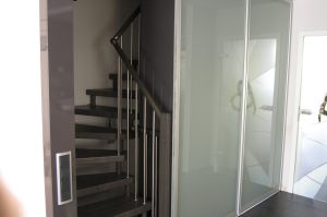 Treppenhaus- Windfang als Hochglanz- Schrank  antrazitfarben   (In der Mitte, Türen grifflos).  Gleittüren mit Mattfolienglasfüllung R+L zum begehen des Treppenhauses.